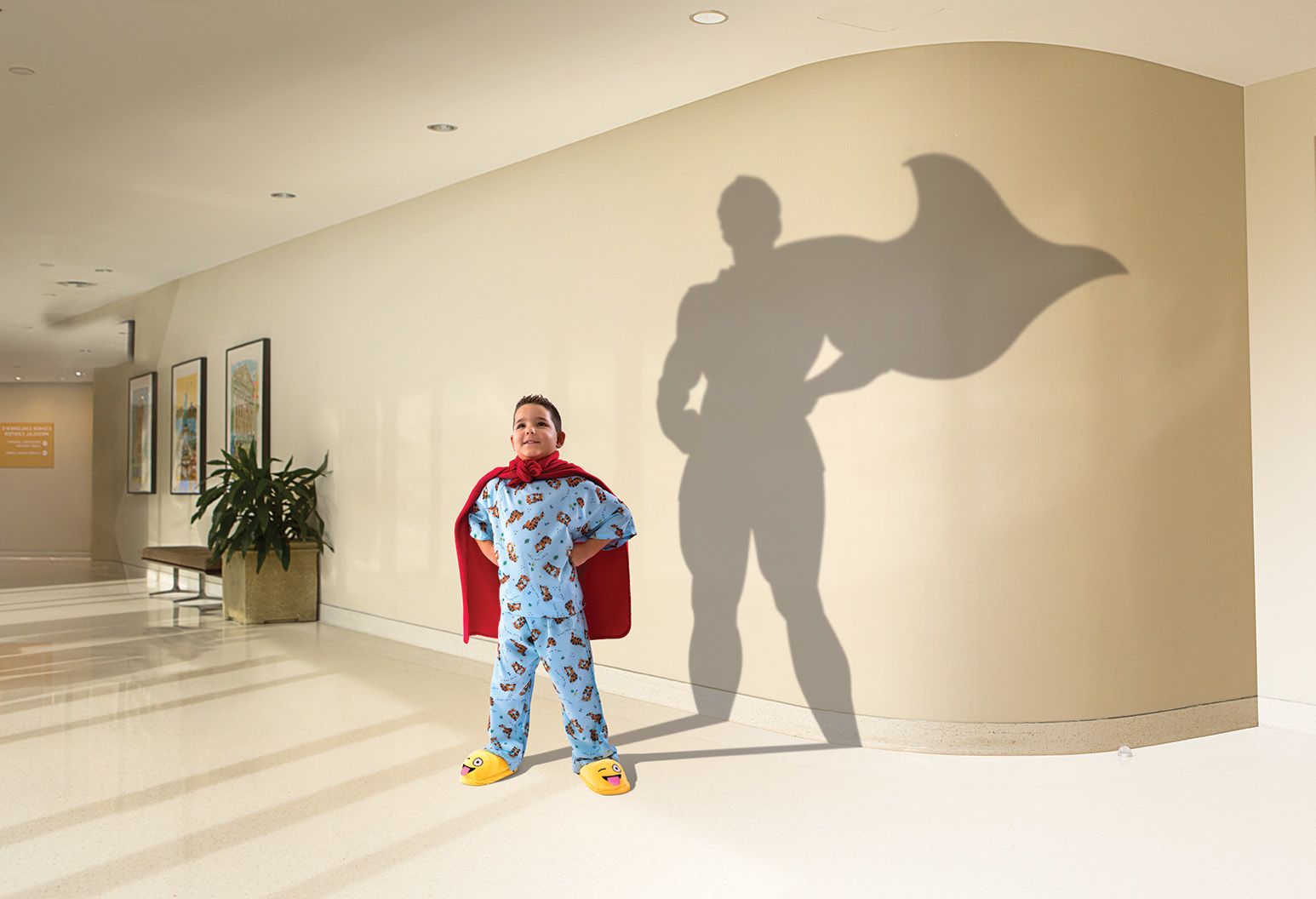 In a hospital room, a five year old by stands tall wearing blue pajamas and his yellow slippers. His shadow is cast on the wall behind him, and the shadow resembles a larger-than-life male super hero with his cape flowing in the wind.